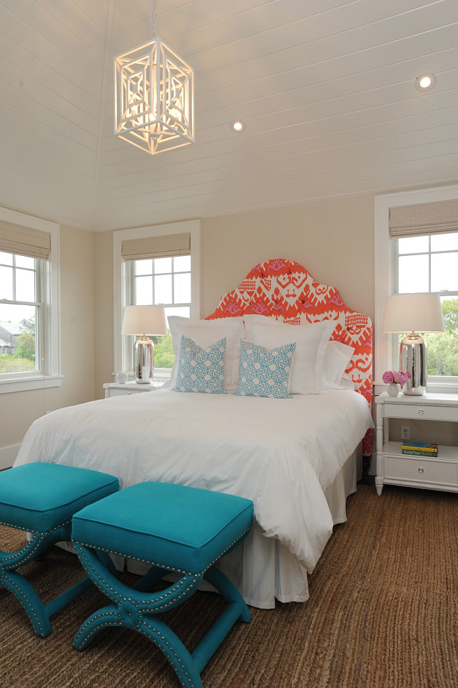 Bedroom. Beach House Bedroom Ideas. Beach House bedroom with tongue and groove, cathedral ceilings adorned with a geometric lantern pendant hung over an orange ikat headboard on bed layered with white bed linens and a pair of blue geometric pillows. The bed is flanked by a pair of white two drawer nightstands topped with mercury glass lamps with a pair of Arteriors Home Tennyson Turquoise Linen Stools at the foot of the bed over hardwood floors layered with a braided jute rug. #Bedroom #BeachHouse #BeachHouseBedroom Nina Liddle Design.