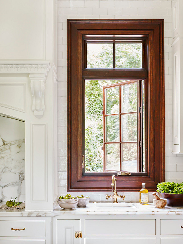 Traditional kitchen kitchen features a cooking nook filled with a curved paneled vent hood with millwork over a marble slab backsplash flanked by spice shelves and a Viking range. A small prep sink stands next to the cooktop paired with a gold vintage faucet placed under a window accented with cherry stained moldings. #kitchen #TraditionalKitchen #window JackBilt Homes.