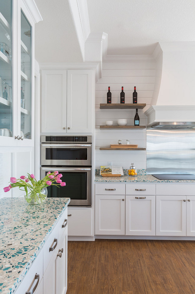 Kitchen Ideas. kitchen features glass-front upper cabinets and white lower cabinets paired with turquoise recycled glass countertops across from a white kitchen hood which is flanked by stacked floating shelves situated over an induction cooktop and a stainless steel cooktop backsplash next to microwave and oven. #KItchen #Ideas #KitchenIdeas Laura U, Inc. 