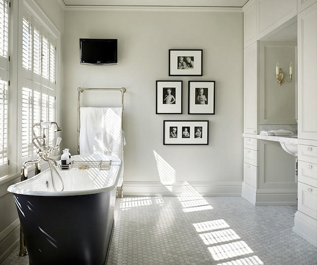 Bathroom. Blacj and White Bathroom. Black and white bathroom with cast iron tub with floor-mount tub filler and bath caddy placed in front of windows covered in plantation shutters over carrara marble hex tile floor. Bathroom features floor to ceiling white built-in cabinets and carrara marble top floating vanity. Bathroom wall with TV, wall-mounted towel warmer and collection of black and white family photos in black gallery frames. Master Bathroom. Toth Construction.