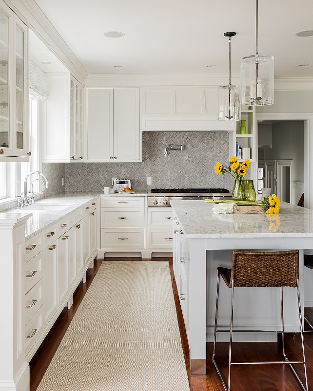 Gray Herringbone Backsplash. Transitional White Kitchen. Transitional White Kitchen with Grey Herringbone Backsplash. This transitional kitchen features white shaker cabinets paired with white marble countertops and a grey marble herringbone tile backsplash. A kitchen sink is paired with a deck mount gooseneck faucet placed under windows flanked by glass front cabinets. A pair of glass column pendants illuminates a marble top island lined with woven barstools. A white kitchen hood stands over a swing arm pot filler and an integrated gas cooktop above pot and pan drawers. #GreyHerringboneBacksplash Jennifer Palumbo.