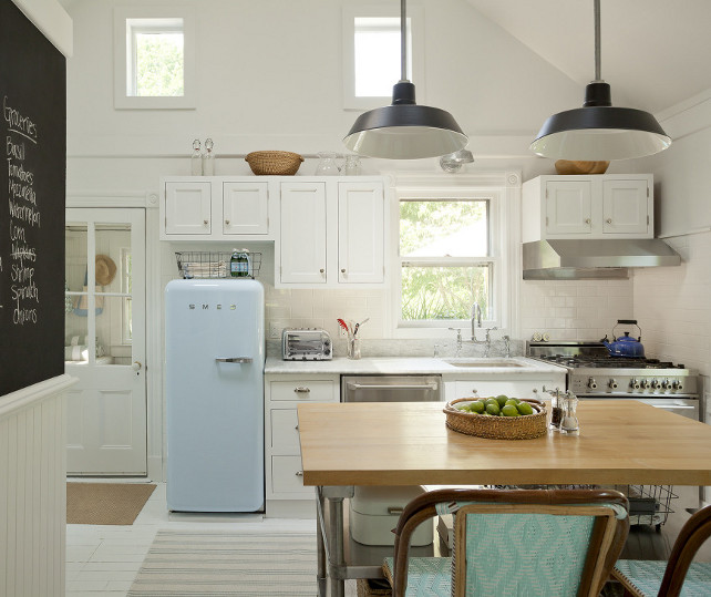 Kitchen - Cottage Kitchen - Cottage. Cottage Kitchen with a blue Smeg Refrigerator. Cottage kitchen features vaulted ceiling adorned with black vintage barn pendants illuminating a freestanding stainless steel island topped with a butcher block countertop lined with turquoise bistro barstools across from a kitchen chalkboard with shelf. White shaker kitchen cabinets are paired with white marble countertops and a white subway tiled backsplash. A small stainless steel stove stands under a stainless steel hood stacked with cabinets beside window above small sink accented with deck-mount bridge faucet alongside a blue Smeg Refrigerator. #Cottage #KitchenCottage #Refrigerator #ColorfulRefrigerator #Smeg #Refrigerator Jenny Wolf Interiors.