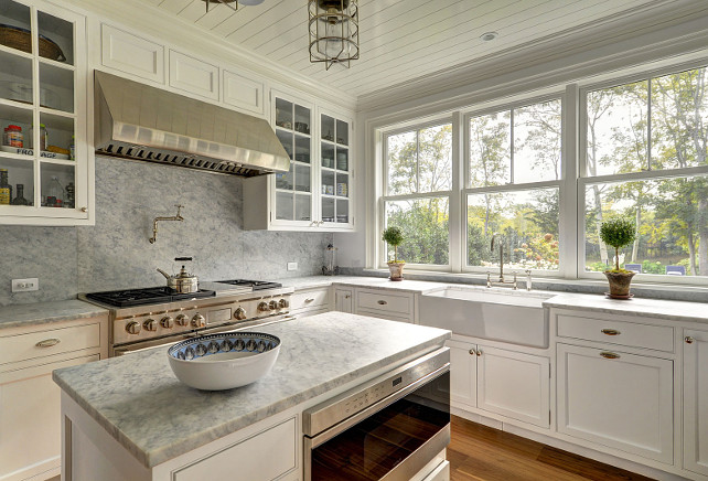 Kitchen with marble slab backsplash. Kitchen with white gray marble slab backsplash. This lovely cottage kitchen features creamy white shaker cabinets paired with gray marble countertops and a gray marble slab backsplash. The kitchen also features beadboard ceiling, a small kitchen island fitted with microwave and a farmhouse sink with deck mount bridge faucet. The range stands under a stainless steel hood flanked by glass-front cabinets. Kitchen lighting are the "Ralph Lauren Crosby Medium Flush Mount In Polished Nickel".#Kitchen #kitchenMarbleSlabBacksplash #SlabBacksplashJohn Hummel and Associates.