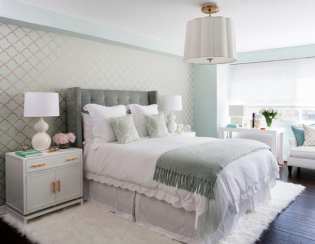 Master Bedroom. Seafoam master bedroom features an accent wall clad in seafoam and gold scallops wallpaper lined with a gray velvet wingback headboard on bed dressed in white scalloped bedding, green Moroccan tiles pillows in Windsor Smith Riad Fabric and a gray fringed throw blanket. A gray tufted headboard is flanked by white nightstands with gold bamboo pulls, Worlds Away Marcus White & Gold Leaf Cabinet, topped with white glass lamps atop a white sheepskin rug lit by a Barbara Barry Simple Scallop Pendant. Master bedroom boasts a bed situated across from a West Elm Parsons Desk placed under windows dressed in white roller shades. #MasterBedroom