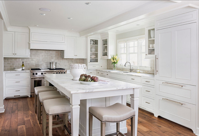 White Kitchen. Classic white kitchen with inset cabinets painted Benjamin Moore Simply White accented with brushed nickel pulls and calcutta countertops with an Encore Silver Ceramic Tiled backsplash. U-shaped kitchen with inset cabinets painted Benjamin Moore Simply White accented with brushed nickel pulls and calcutta countertops with an Encore Silver Ceramic Tiled backsplash. A dual stainless steel range stands at the far end of the island finished with a paneled range hood. The kitchen features a stainless steel undermount sink with gooseneck faucet situated below a triple sash window with glass front cabinets either side next to a cabinet front refrigerator. The long kitchen island is lined with Hickory Chair Madigan Counter Stools which are tucked below the countertop overhang. #kitchen #Whitekitchen #classickitchen #benjaminMooreSimplyWhite 